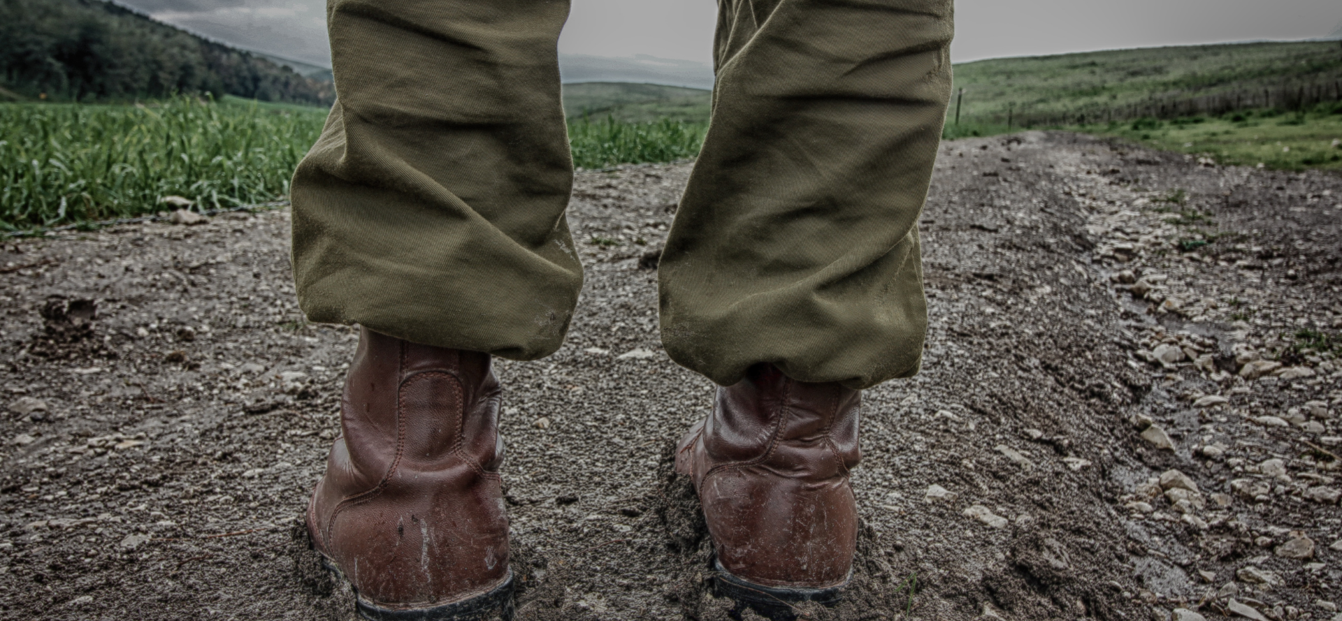 Lower legs of person wearing military-style pants and boots, standing on a path in a rural area